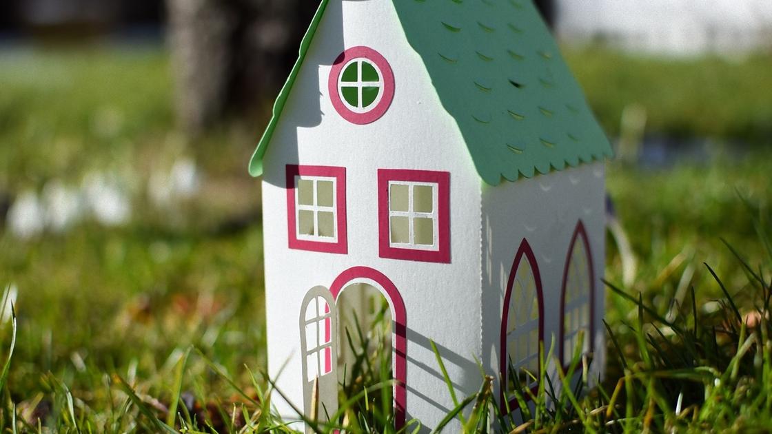 Бумажный домик с зеленой крышей, красными украшениями на окнах и открытой дверью стоит в траве